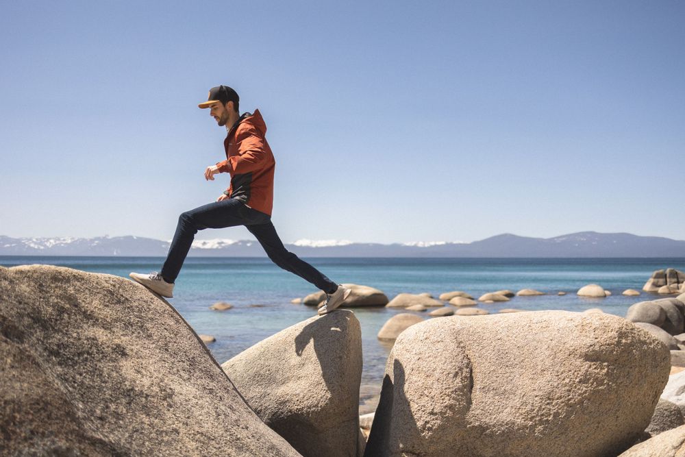 An adventurer jumps between large rocks at Lake Tahoe with mountains in the background.