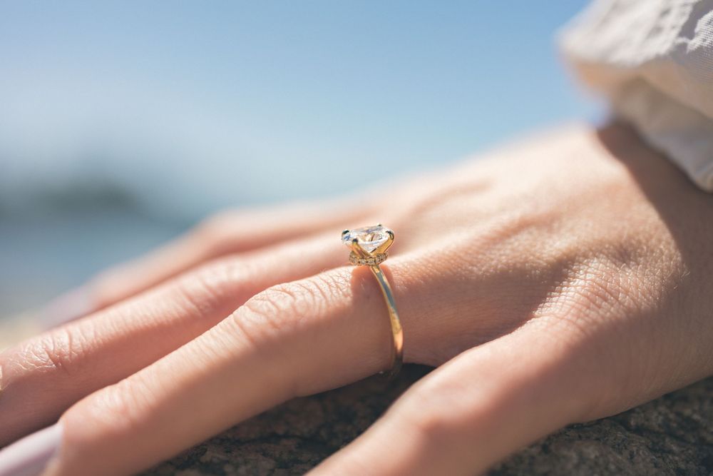 Close-up of a delicate diamond engagement ring on a woman's hand against a blurred beach background.