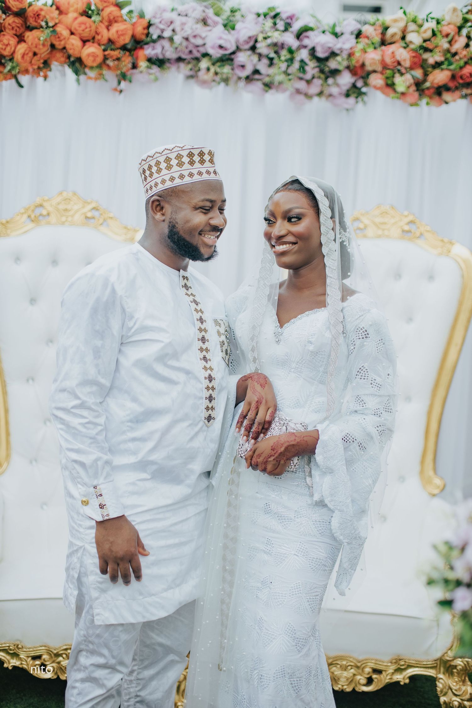 A wedding couple in matching white traditional attire poses against a floral backdrop.