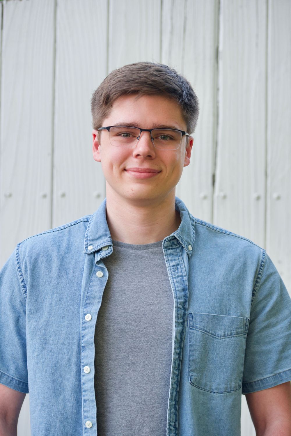 Casual portrait of person in light denim shirt and glasses against white wall background.