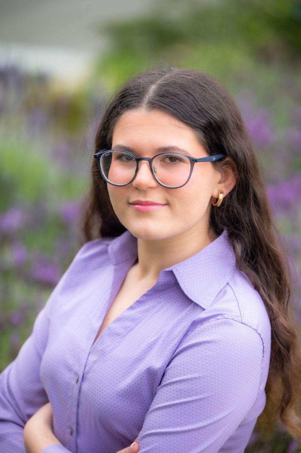 Professional headshot of person in lavender shirt with glasses against floral garden background.
