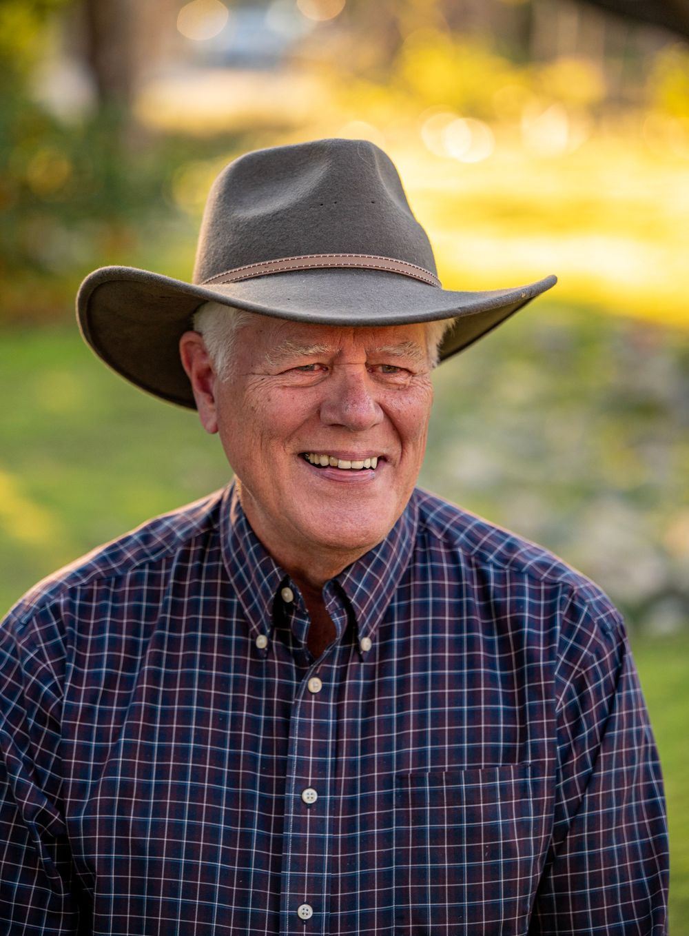 Senior man in cowboy hat and plaid shirt smiling outdoors with golden bokeh background.