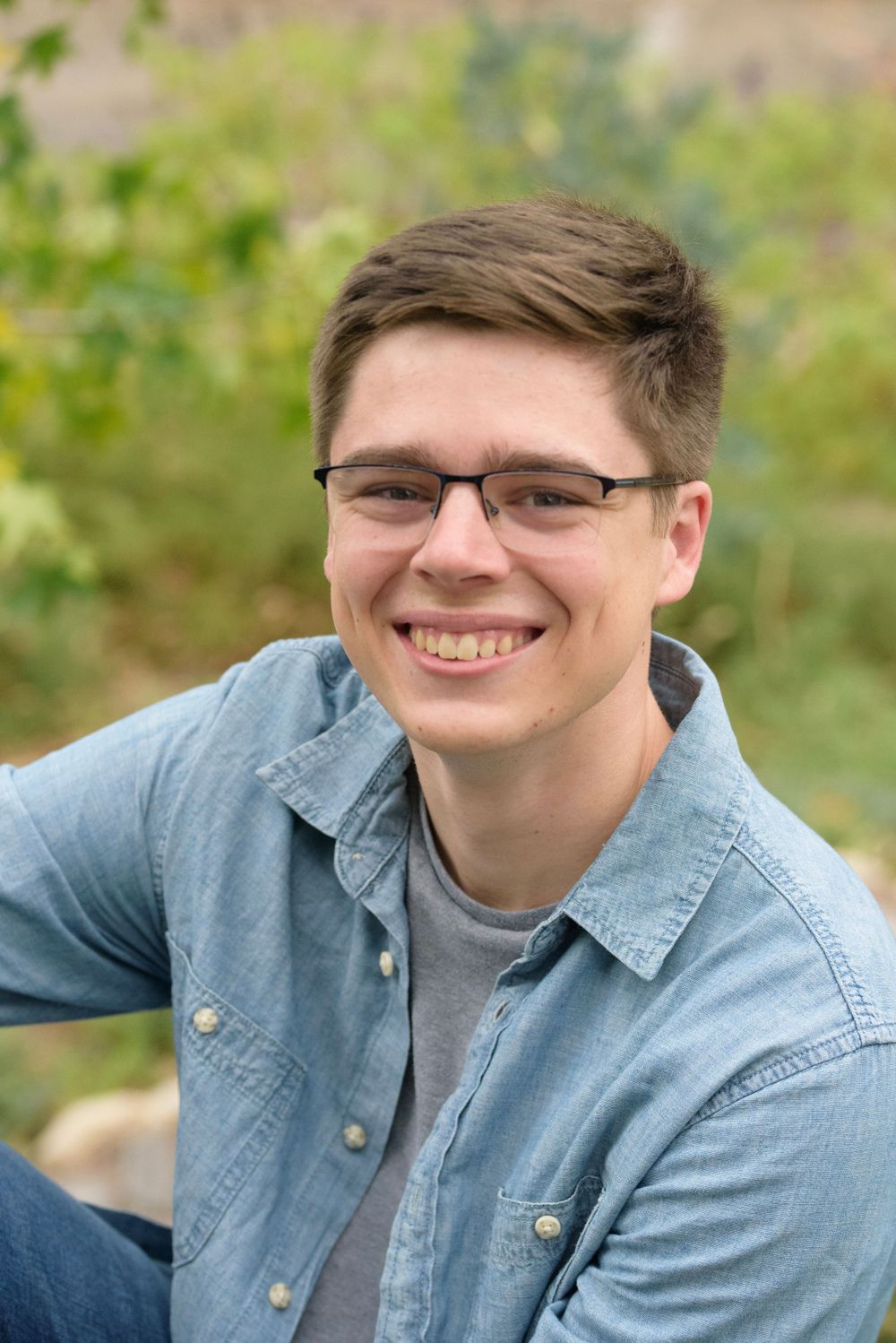 Professional headshot of someone in casual denim shirt and glasses against green outdoor background.