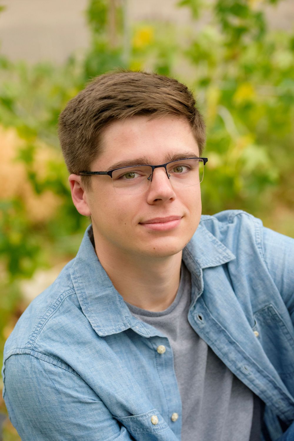 Casual portrait of someone in glasses and denim shirt against green foliage.