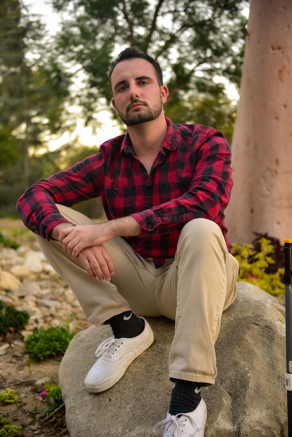 Casual outdoor portrait of person in red flannel and khakis sitting on rock with scenic background.