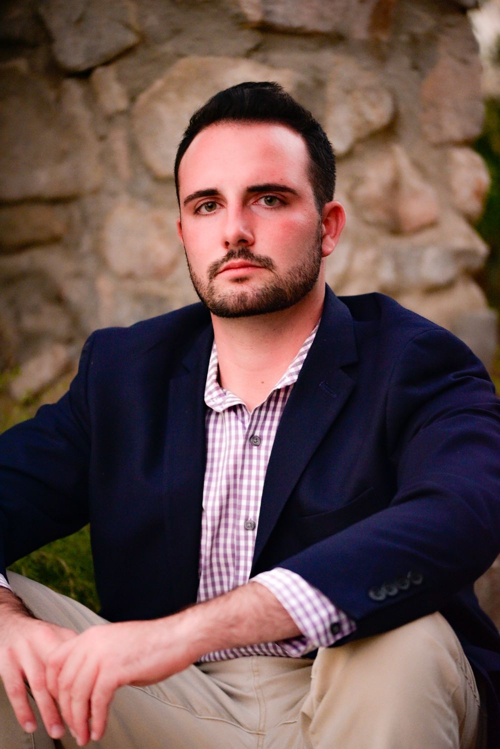 Professional portrait of person in navy blazer and checkered shirt against stone wall background.