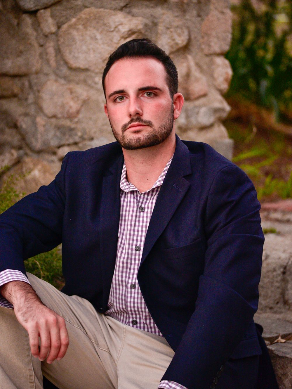 Portrait of person in navy blazer and gingham shirt posed against rustic stone wall.