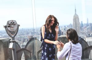 Proposal scene at Top of the Rock observation deck with iconic NYC cityscape and vintage viewfinder.