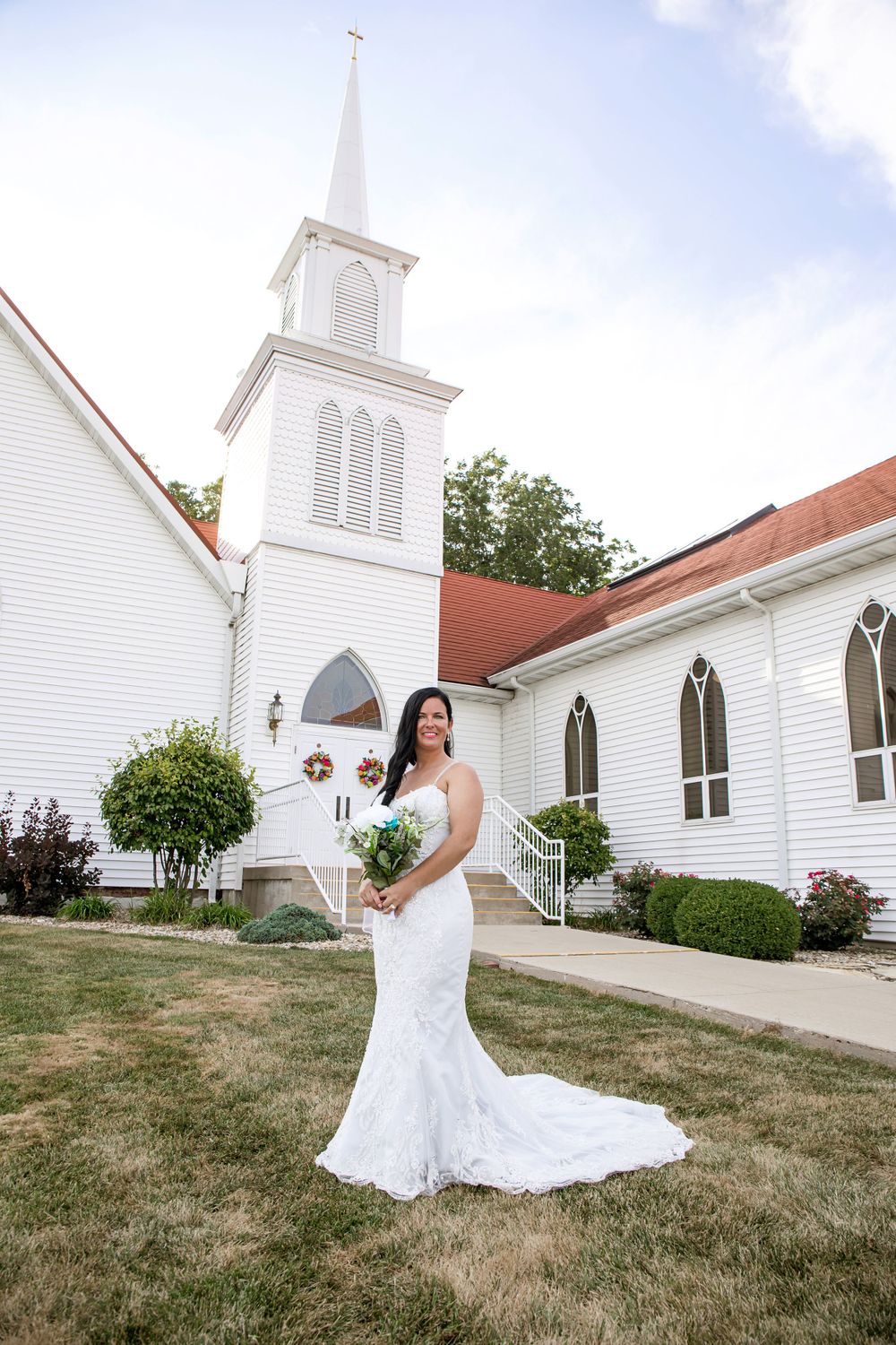 A white church with a tall steeple serves as backdrop for outdoor bridal portraits.