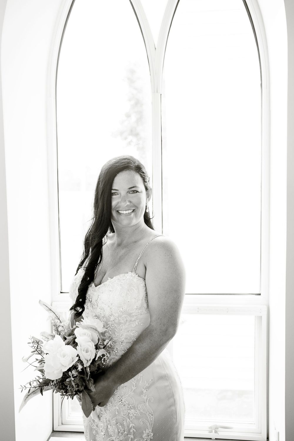 A bridal portrait showing a lace dress and bouquet against arched windows in black and white.