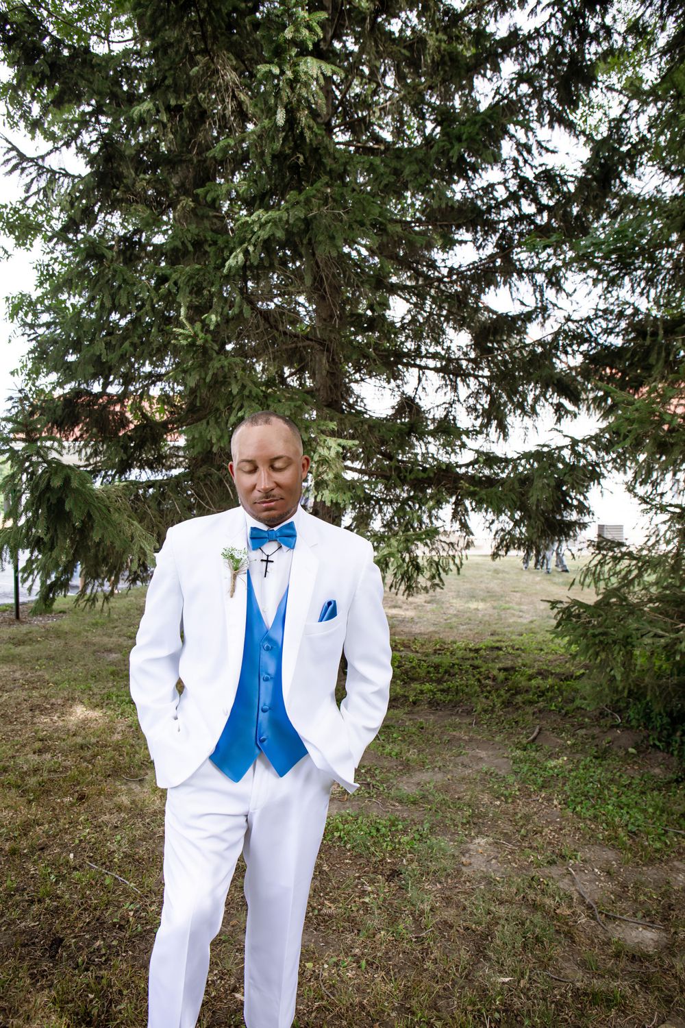 A dapper groom in white tuxedo with bright blue vest and bowtie poses against evergreen trees.
