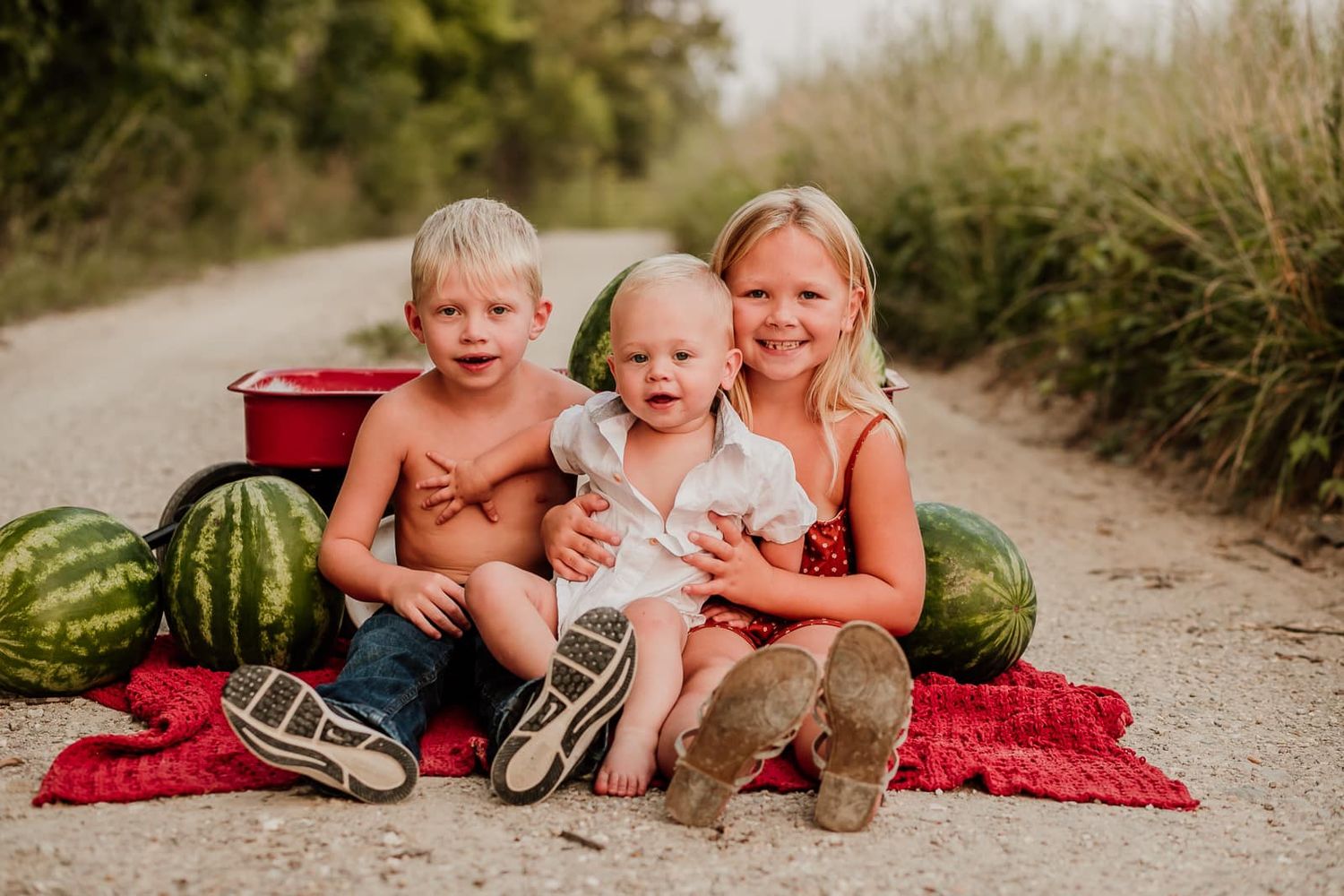 Three children sitting on a red blanket with watermelons on a country road.