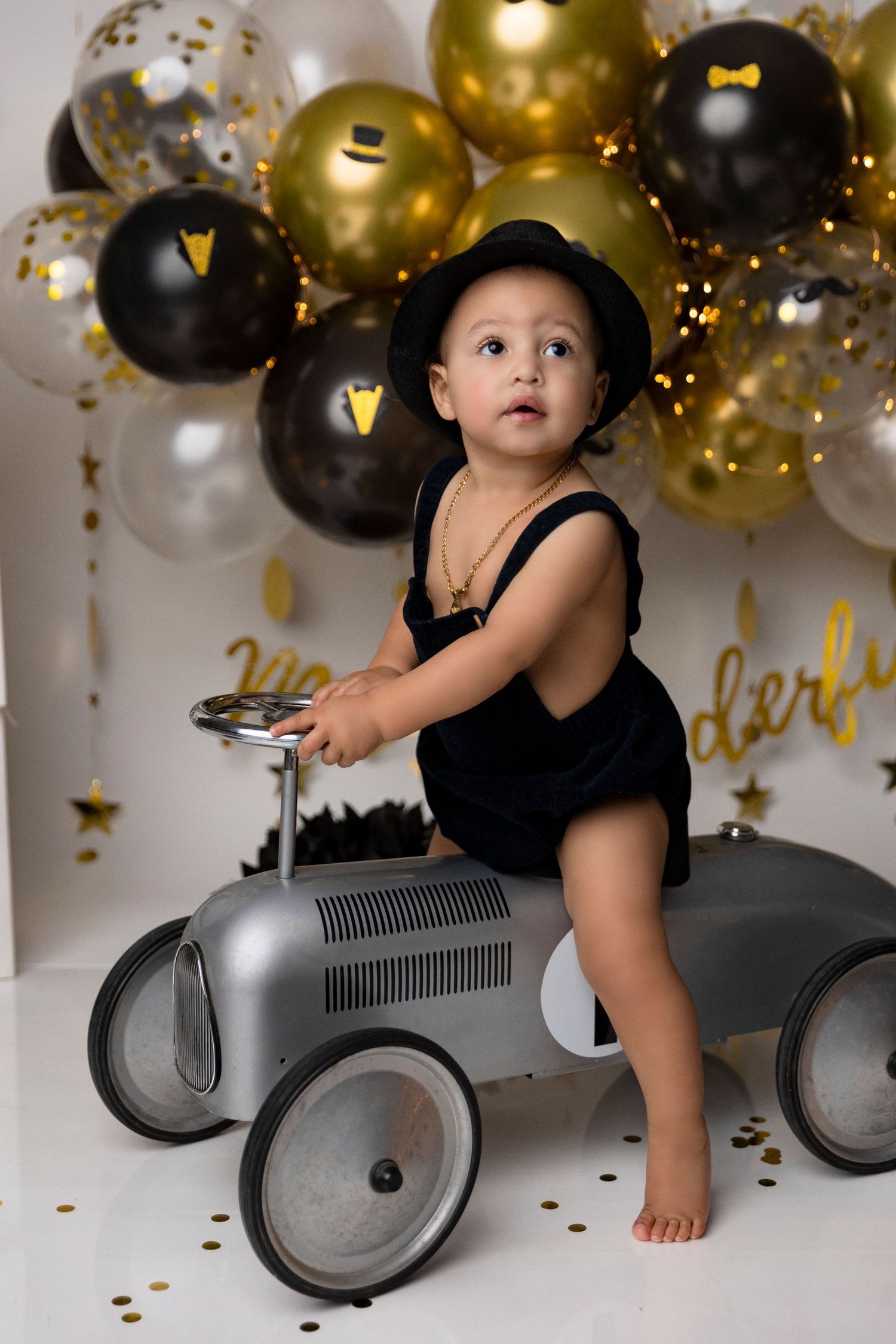 Baby sitting in vintage-style toy car with gold and black balloons in background.