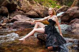 Model in black dress poses artistically on rocky waterfall setting.