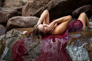 Dramatic pose with burgundy fabric flowing over wet rocks near stream.