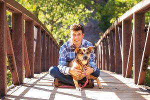 Person in blue plaid shirt sitting with small brown dog on wooden bridge outdoors.