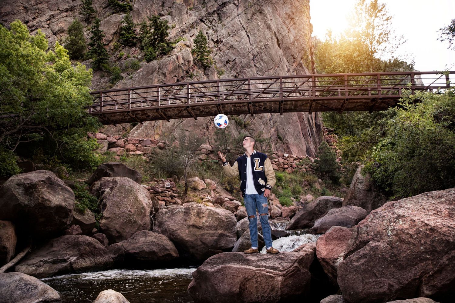A figure stands on rocky terrain beneath a wooden footbridge with dramatic sunlit cliffs in the background.