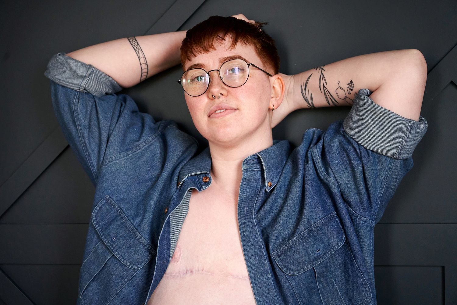 A casual portrait against a dark wall showing denim jacket and glasses.