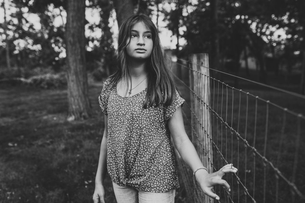 A moody black and white portrait of someone in a patterned top standing by a wire fence in nature.