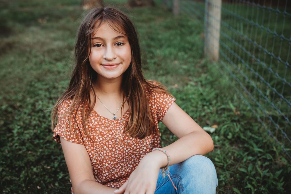 Someone in a floral print top and jeans smiles while sitting in the grass near a fence.
