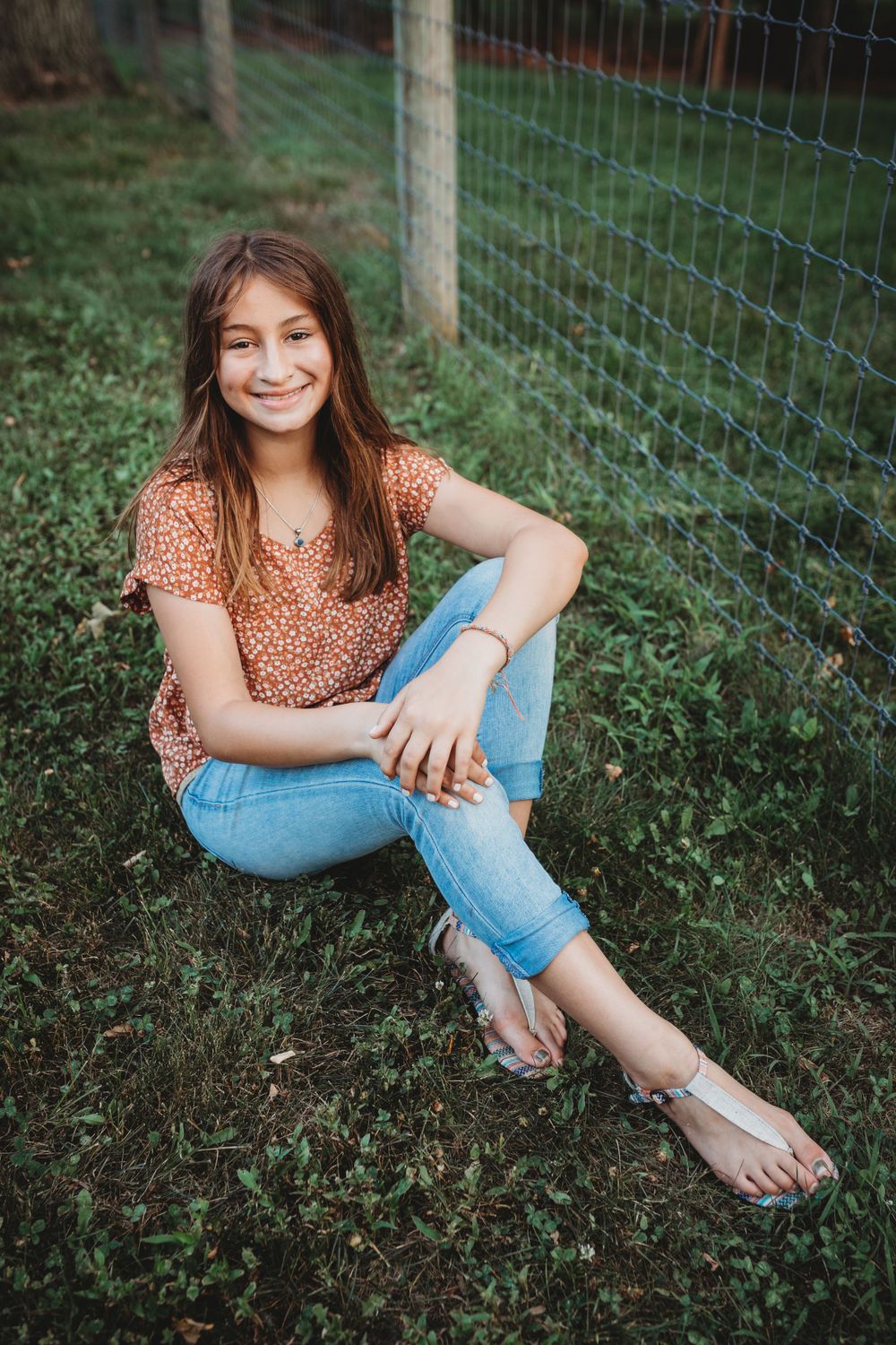 A relaxed outdoor portrait of someone wearing a floral top and jeans sitting by a wire fence.