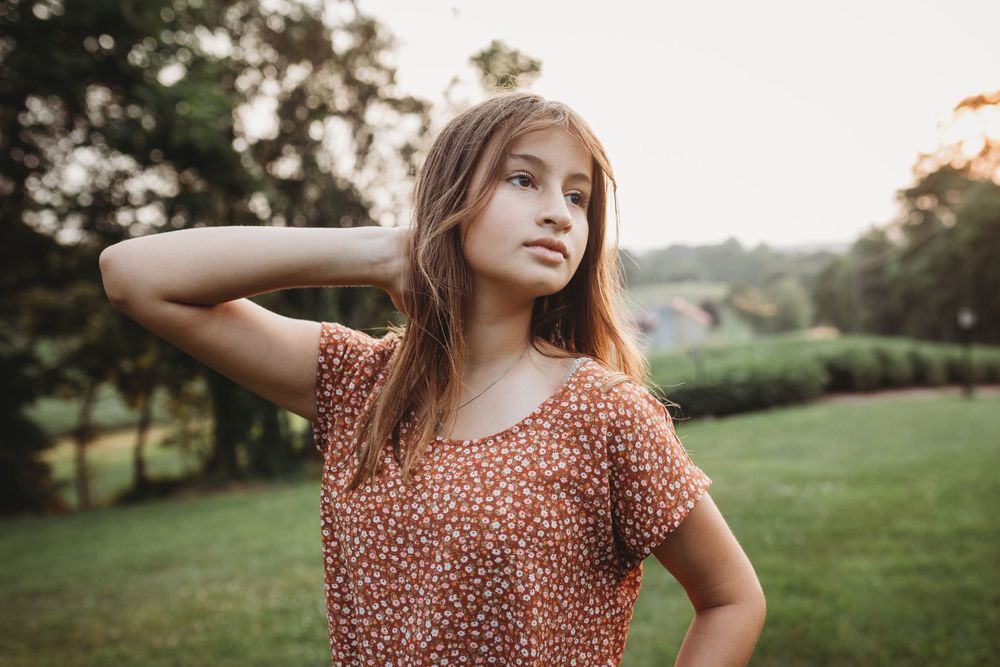 Someone poses thoughtfully in a rust-colored patterned top against a sunset landscape.