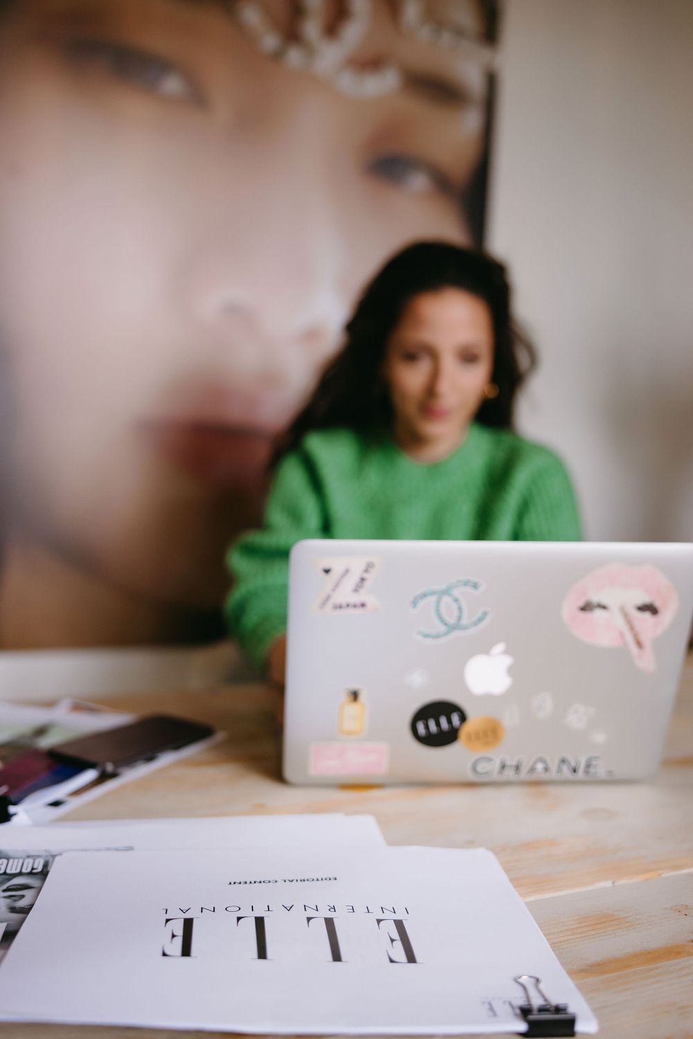 Person in green sweater working at desk with laptop decorated with fashion stickers.