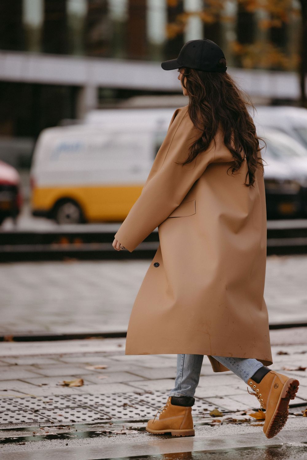 Long camel coat styled with baseball cap and boots on wet pavement.