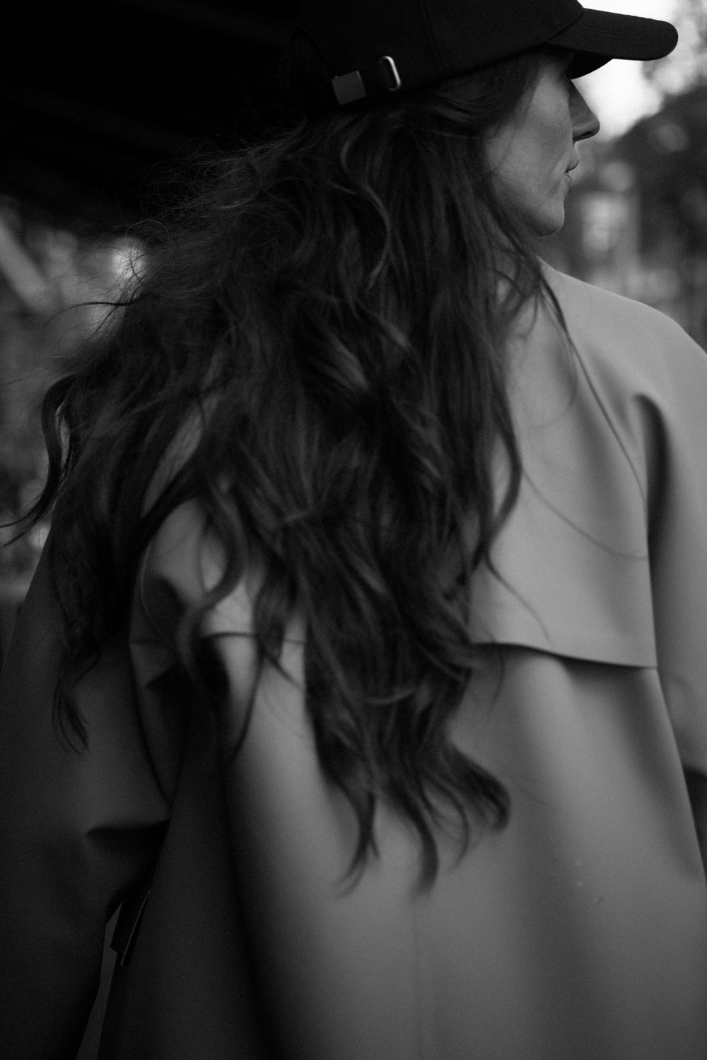 Artistic black and white portrait showing long wavy dark hair and baseball cap in profile against blurred background.