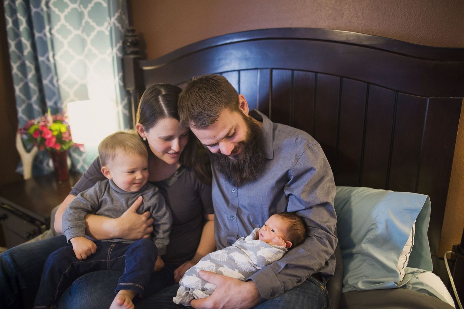 Two caregivers share a tender moment with babies while sitting on a bed near a window.