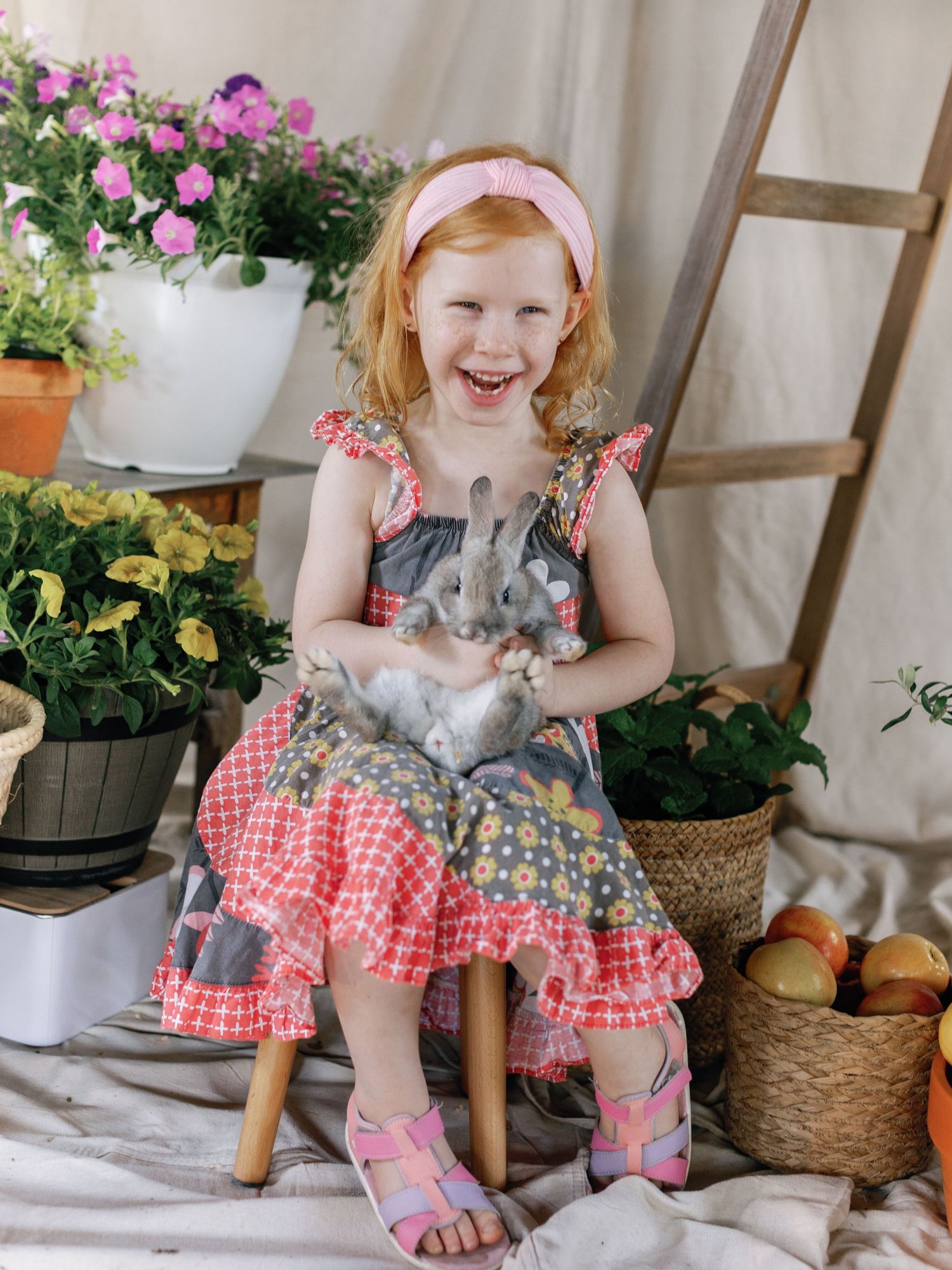 Young child in gingham dress holding rabbits while sitting among potted flowers with a bright smile.