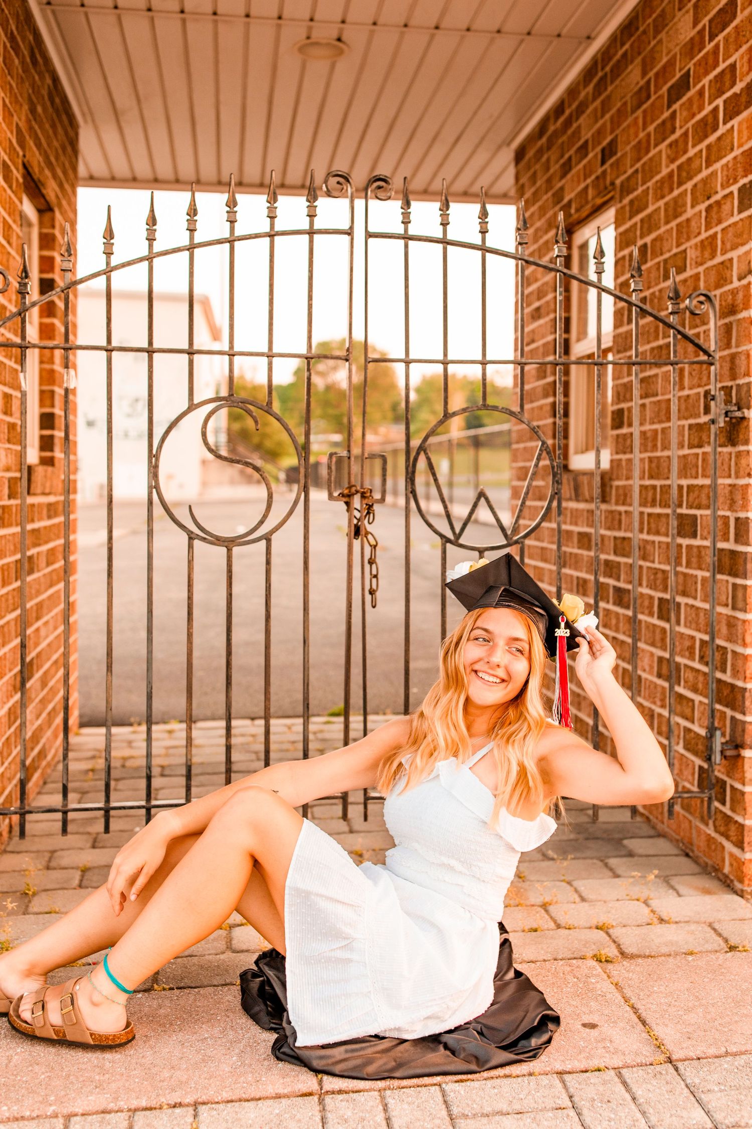 Smiling graduate in white dress and cap sits against ornate iron gate at brick building.