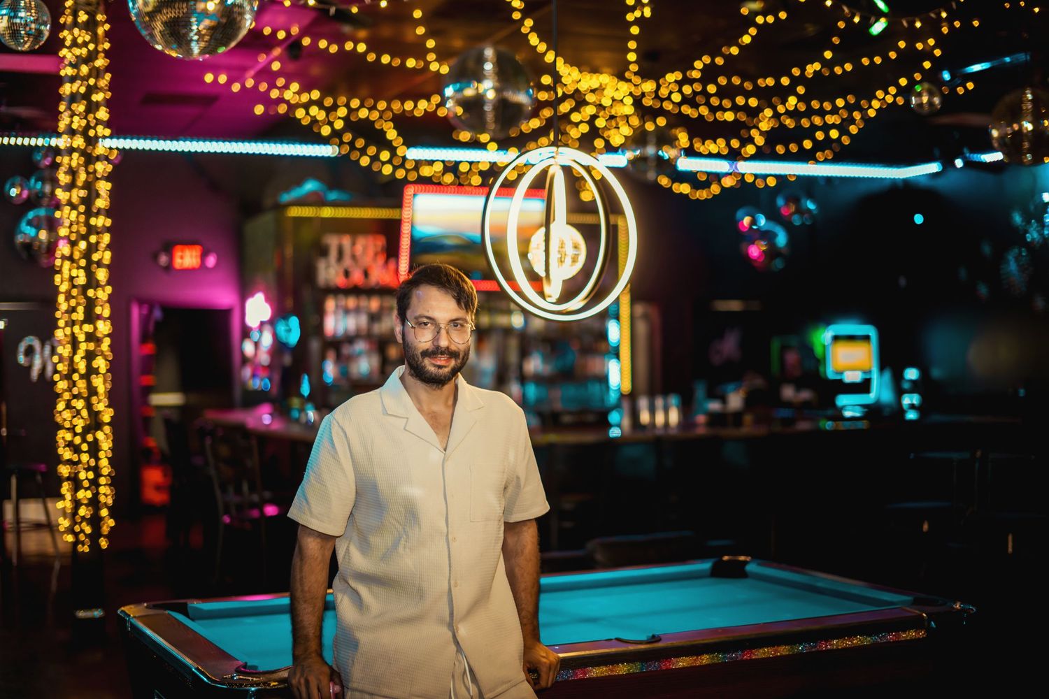 Man in white shirt standing in bar with colorful string lights and pool table.