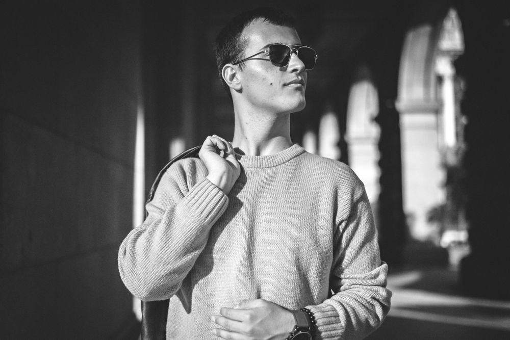 Person in sunglasses and light sweater poses in architectural hallway with dramatic lighting.