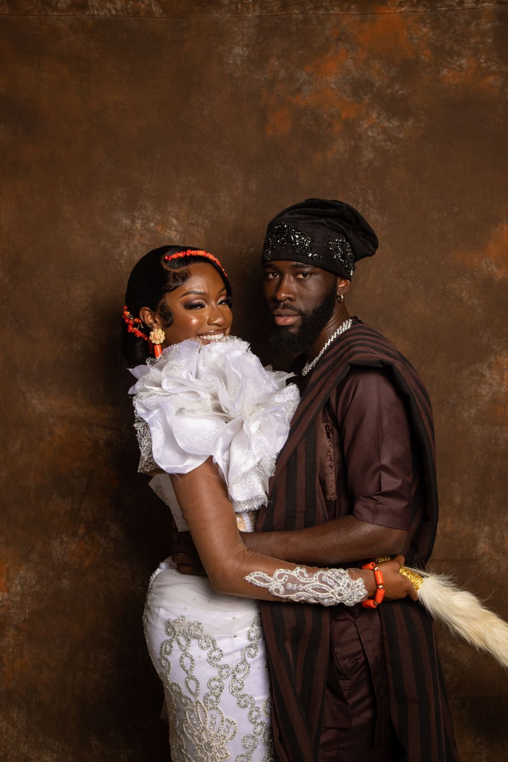 Formal portrait of couple in traditional dress embracing against rustic brown backdrop.