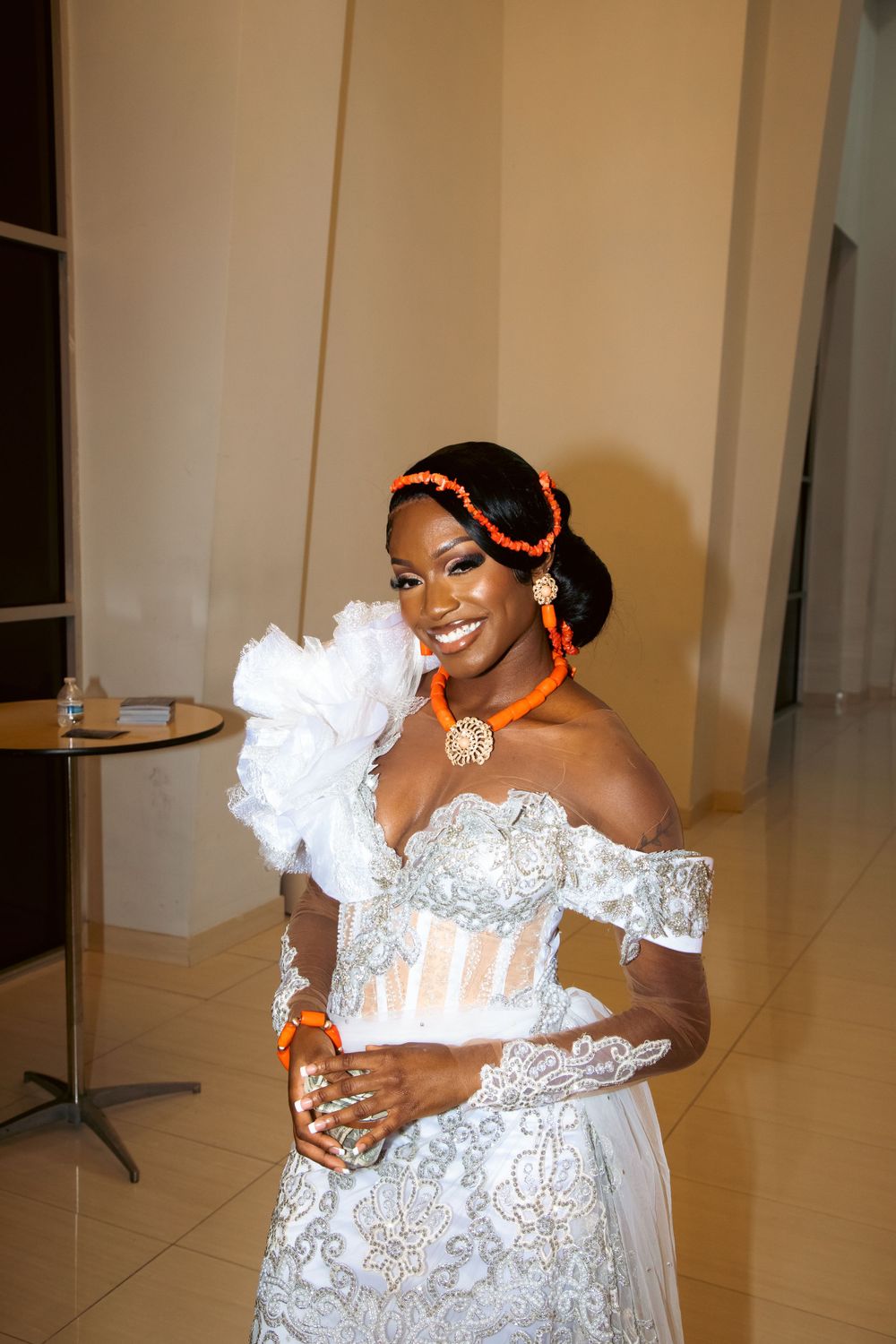 Woman in an ornate white lace dress with orange accessories poses elegantly at a formal event.