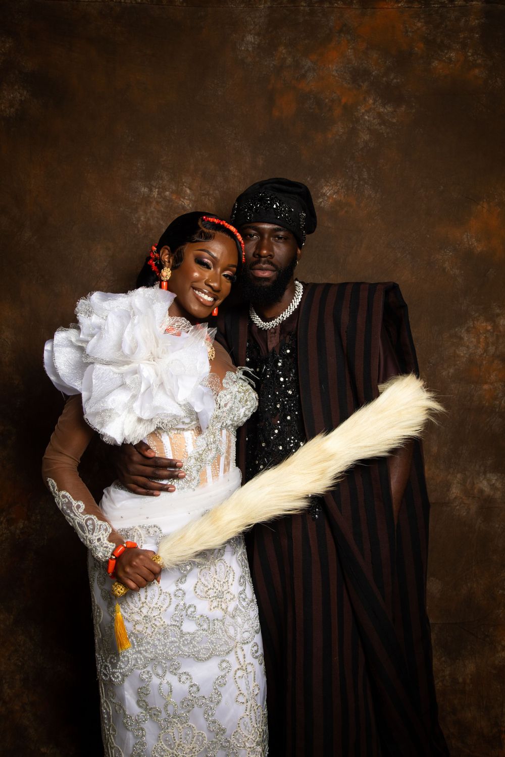 Couple poses in traditional wedding attire against brown backdrop with decorative fan prop.