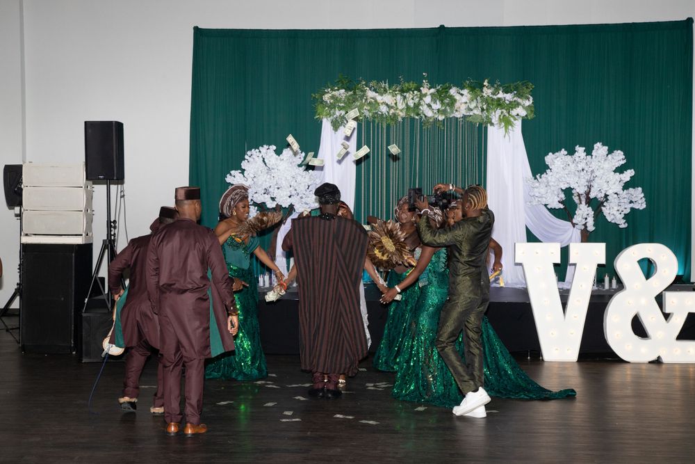 Wedding guests dancing in traditional attire in front of a green backdrop with decorative white elements.
