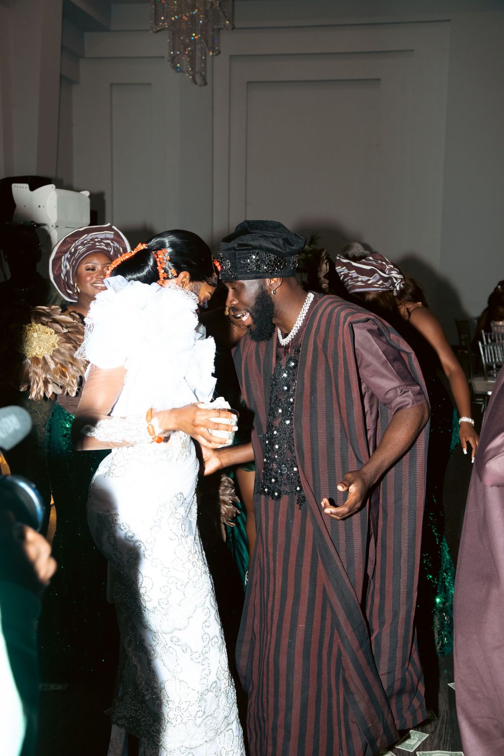 A dancing couple in traditional attire enjoy a celebration, with one in white lace and another in striped formal wear.