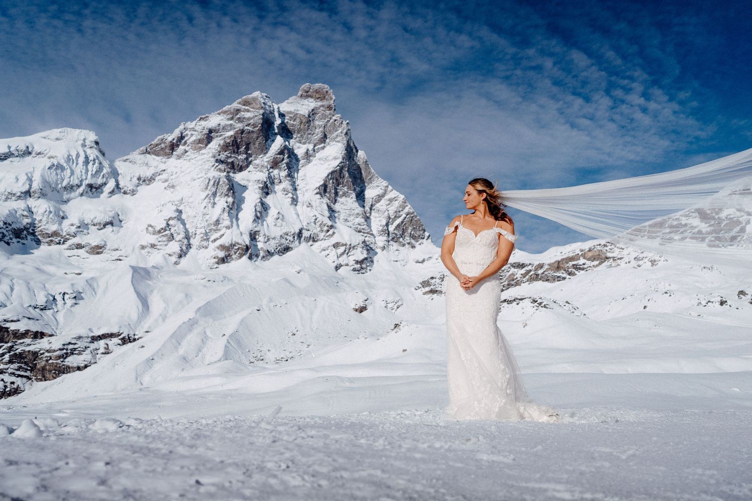A bride in a white gown poses in a snowy mountain landscape with dramatic peaks.