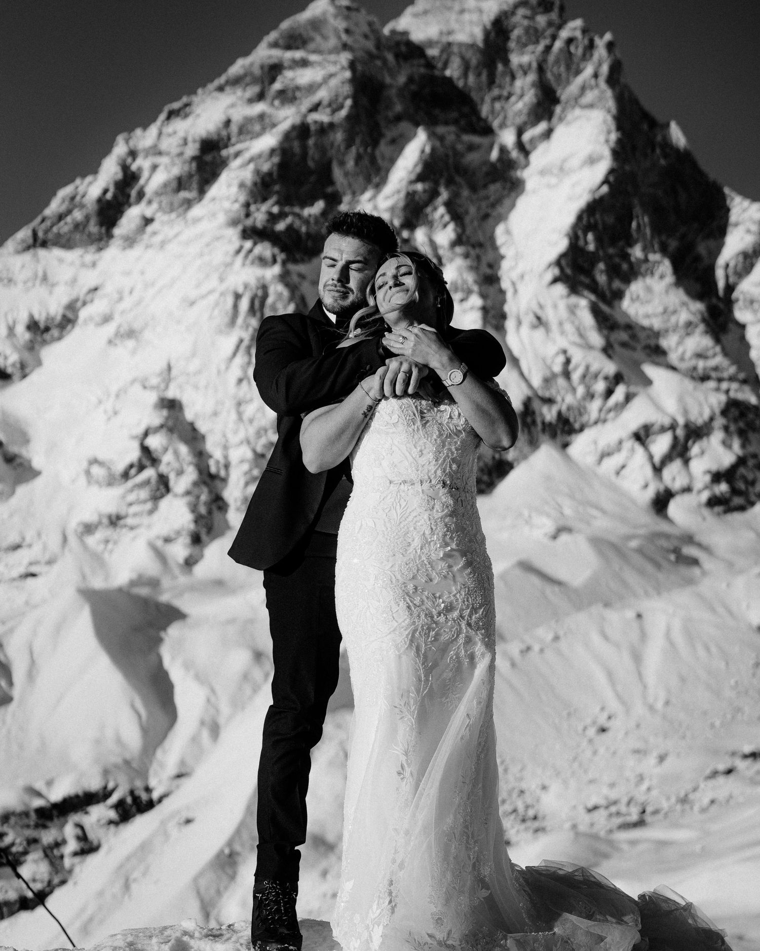 A romantic winter wedding photo showcasing a couple embracing in front of a snow-covered mountain peak.