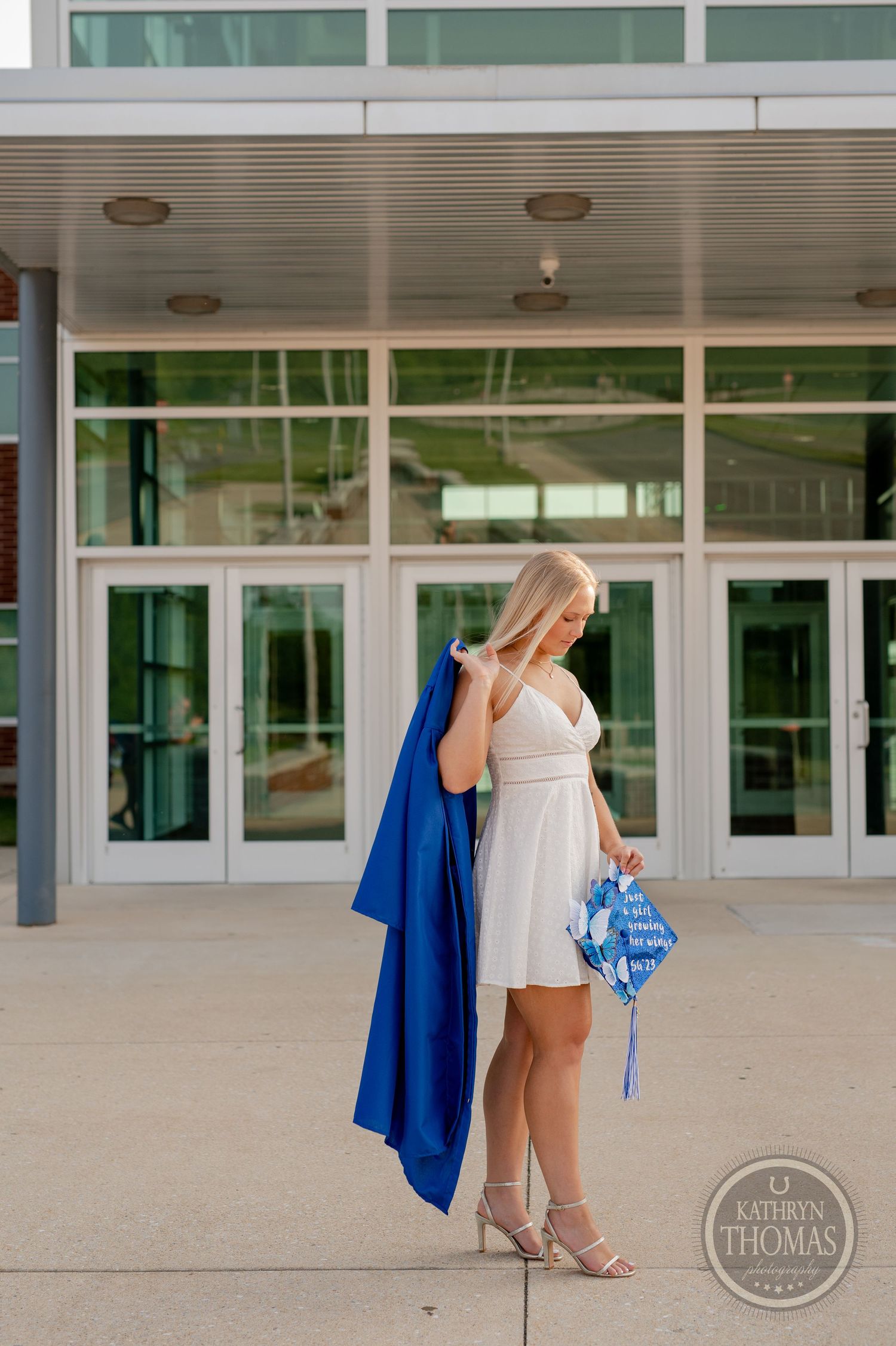 Graduate in white dress and blue graduation gown poses elegantly at school entrance.