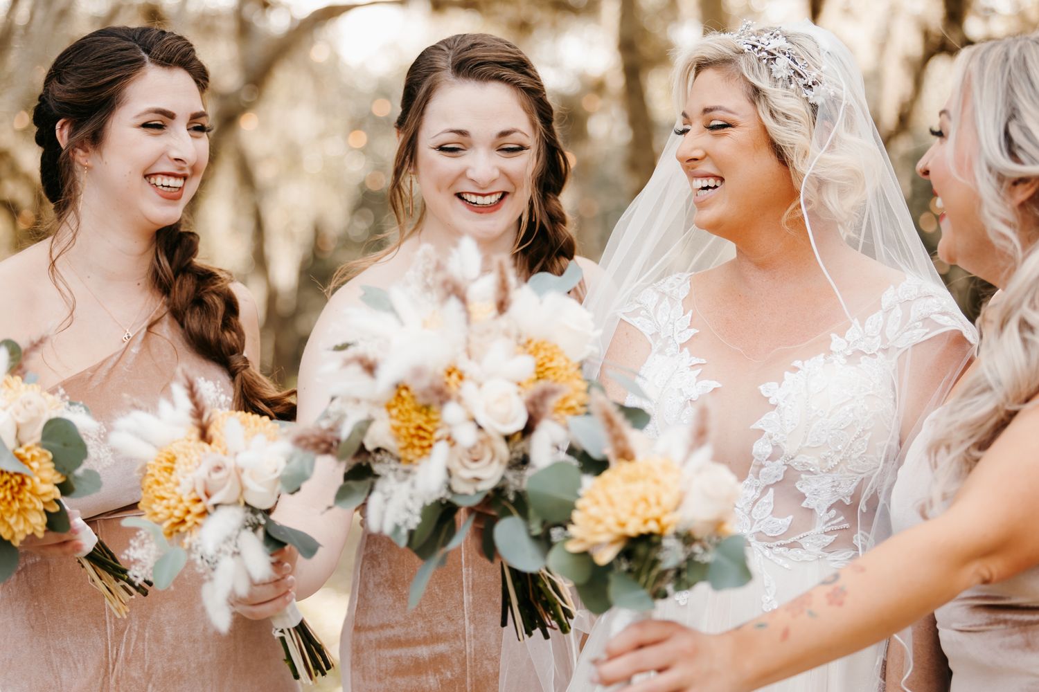 Laughing bride in lace gown with bridesmaids in blush dresses holding white and yellow floral bouquets.