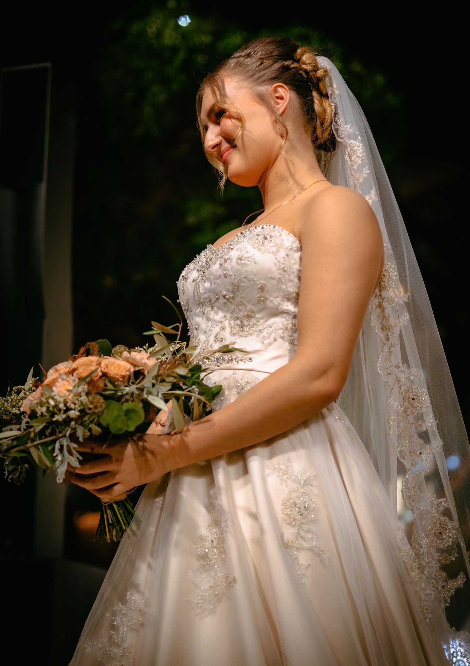 Smiling bride in a strapless lace wedding gown holding a peach floral bouquet at night.