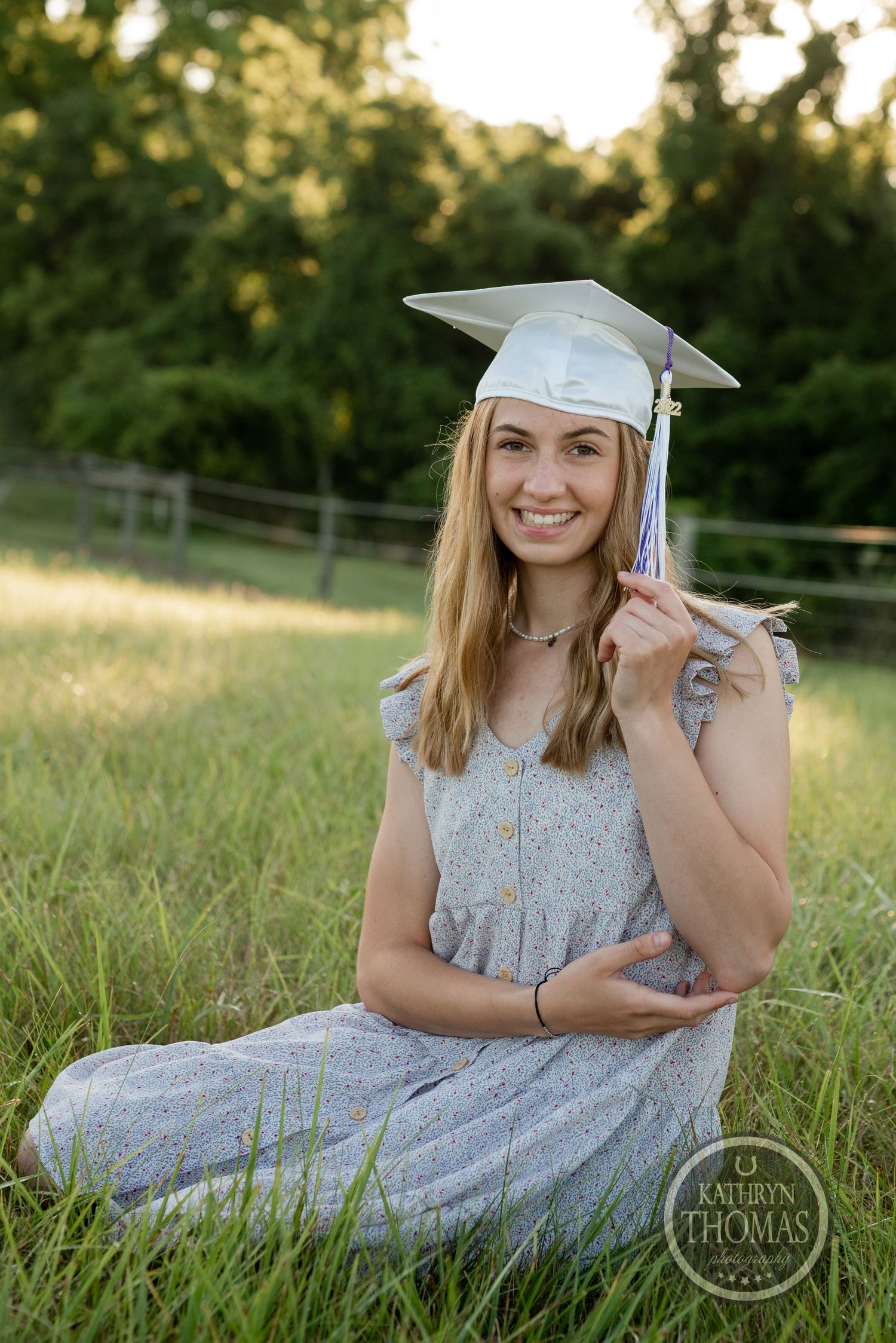 Graduate in grey dress and white cap poses playfully in grassy field during sunset photoshoot.