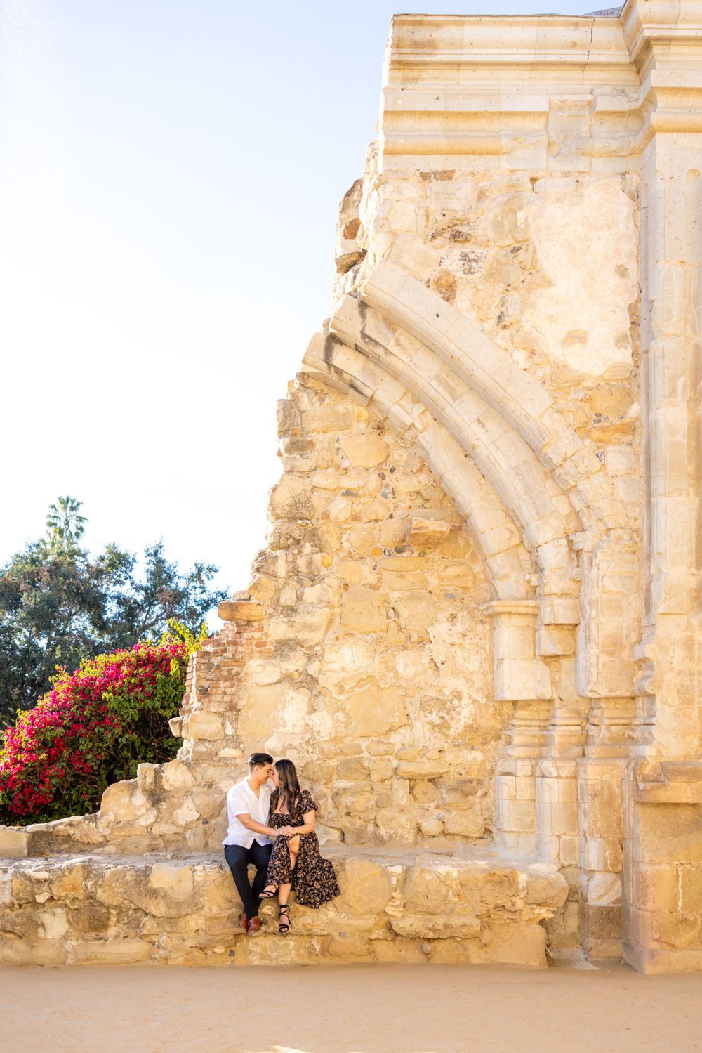 Couple sitting on stone wall by Mediterranean architecture with bright bougainvillea flowers.