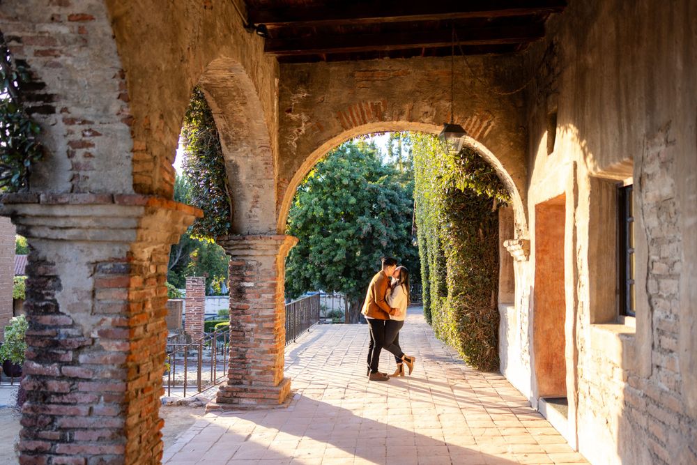 Silhouetted figures embrace under arched mission walkway with green foliage.