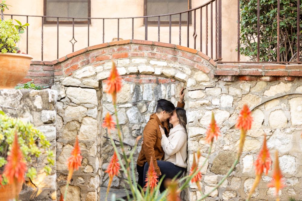Couple embraces by stone wall with orange flowering plants in foreground.