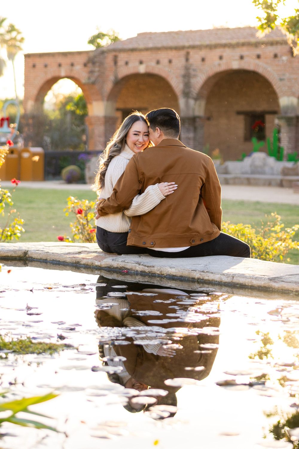 Couple sitting by reflecting pool with historic mission architecture in background at sunset.
