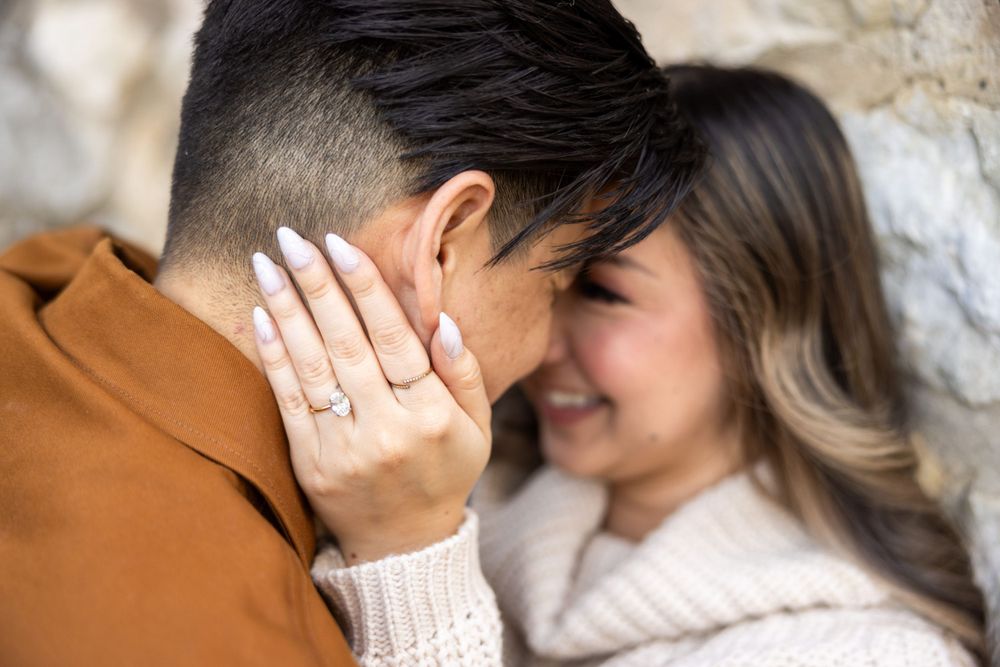 Close up moment between couple showing engagement ring against stone wall background.